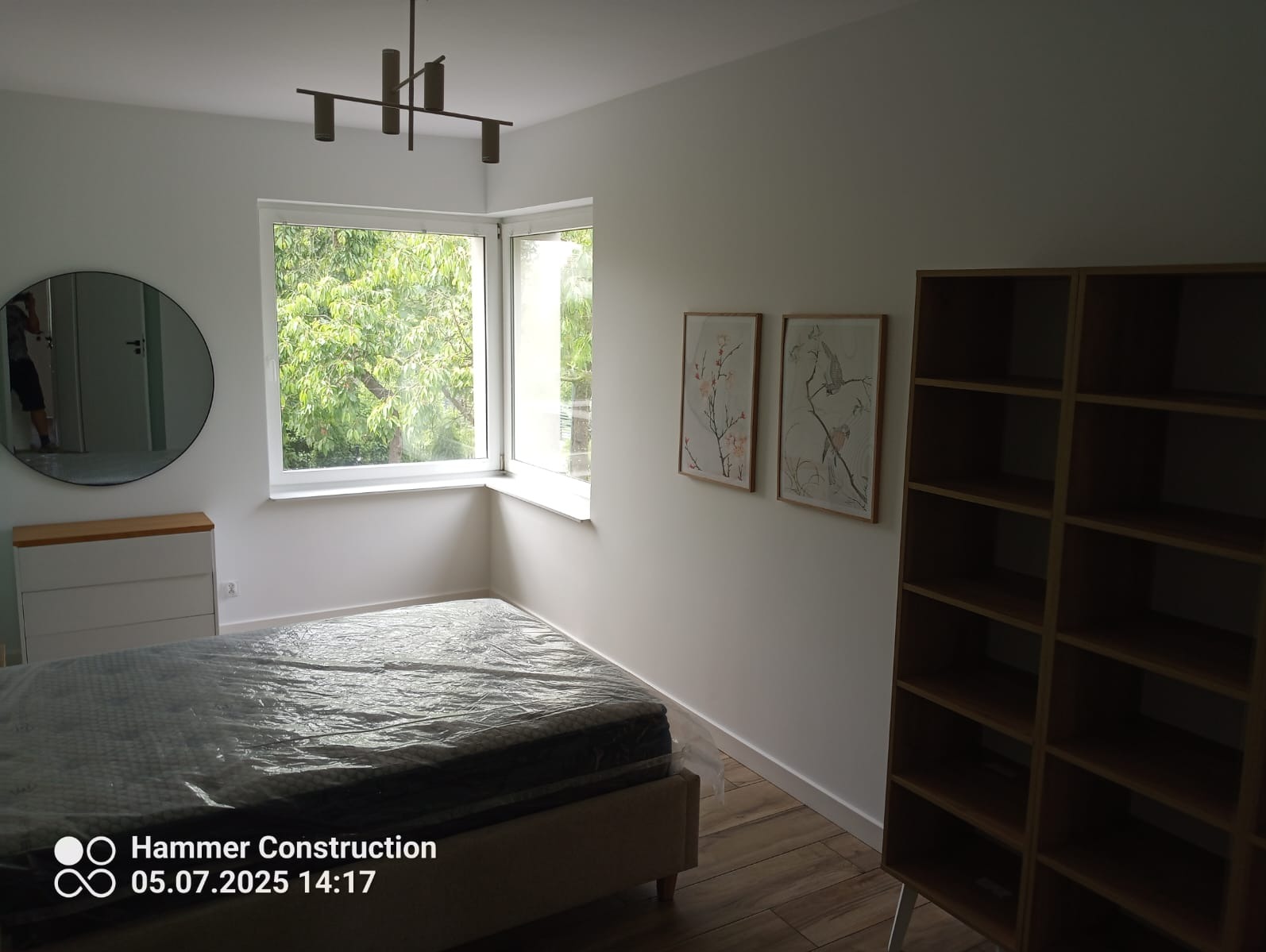 Jasne wnętrze sypialni po remoncie: łóżko przy oknie, okrągłe lustro, komoda, regał i grafiki na ścianie. Podłoga z paneli. Nowoczesna lampa sufitowa.