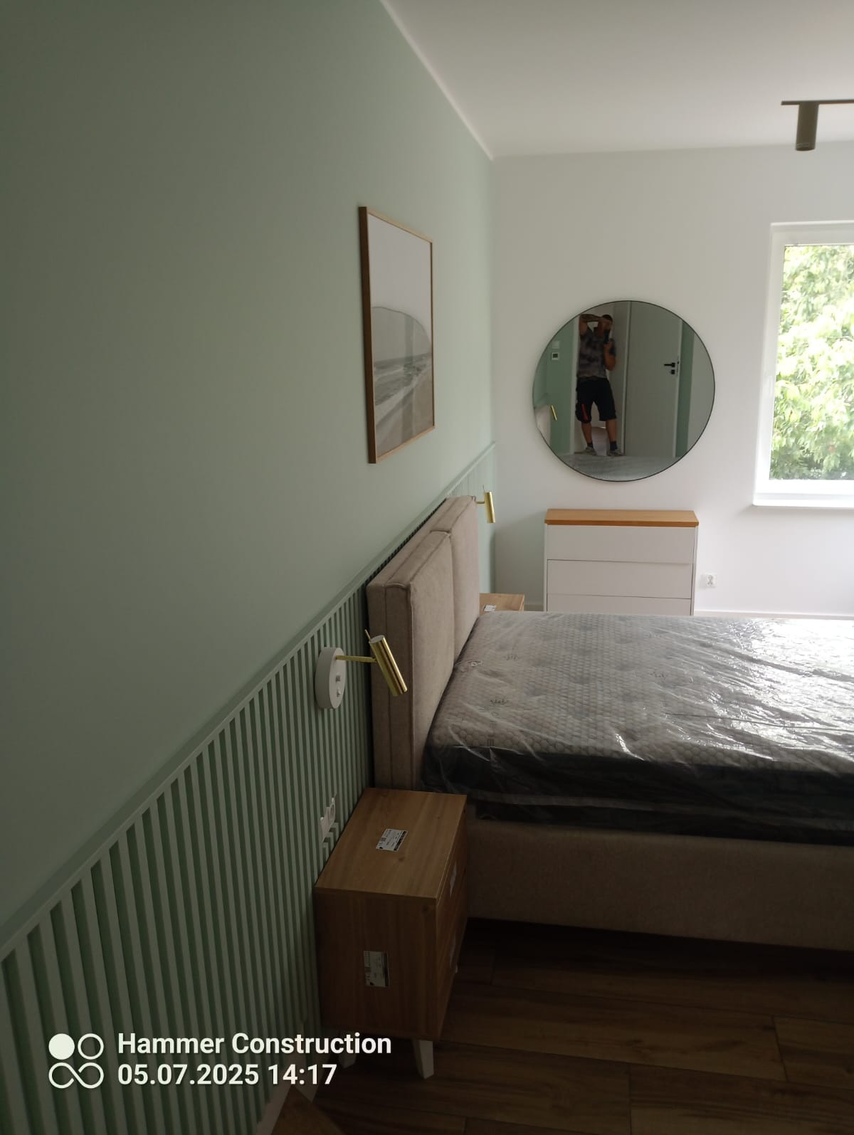 Sypialnia po remoncie: pistacjowa ściana z lameli, łóżko z beżowym zagłówkiem, okrągłe lustro odbija fotografa. Nowoczesny design.