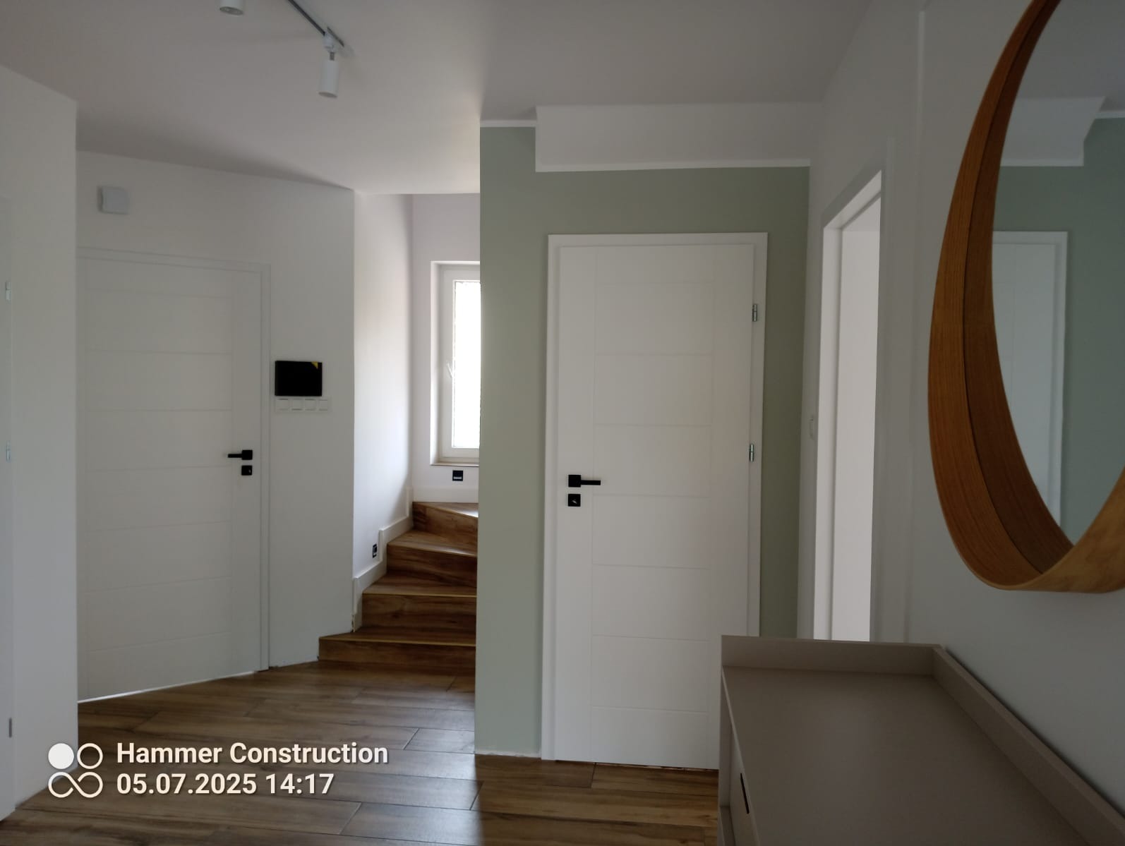 Nowoczesny przedpokój z drewnianymi schodami, białymi drzwiami i podłogą. Uwagę przykuwa duże, okrągłe lustro z drewnianą ramą. Minimalistyczne oświetlenie sufitowe.