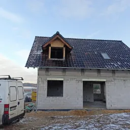 Budowa domu jednorodzinnego w Zielonej Górze z częściowo pokrytym dachem z dachówki ceramicznej, widoczne okno dachowe i konstrukcja lukarny, obok zaparkowany bus firmy dekarskiej.