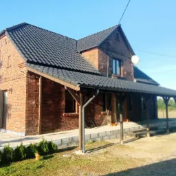 Dom z czerwonej cegły z nowym, ciemnoszarym dachem i zadaszonym tarasem, widoczne rynny i obróbki blacharskie, w tle błękitne niebo i zieleń drzew.