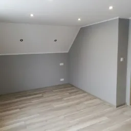 Pomalowane na szaro wnętrze pokoju na poddaszu z panelami podłogowymi imitującymi drewno, widoczne gładkie ściany, biały sufit z oświetleniem punktowym i gniazdka elektryczne.