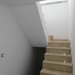 Klatka schodowa z marmurowymi stopniami, białe ściany, widoczne niedoskonałości wykończenia w postaci niezamaskowanych puszek elektrycznych.