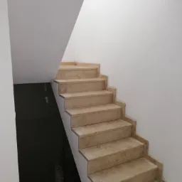 Wykończone schody z marmuru beżowego w jasnym, minimalistycznym wnętrzu. Widoczne białe ściany i sufit. W tle ciemne, niezagospodarowane pomieszczenie.