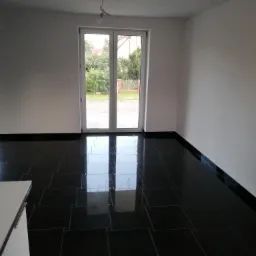 Puste, nowo wykończone wnętrze z błyszczącą, czarną podłogą z płytek, białymi ścianami i dwuskrzydłowymi drzwiami balkonowymi z widokiem na zieleń ogrodu.