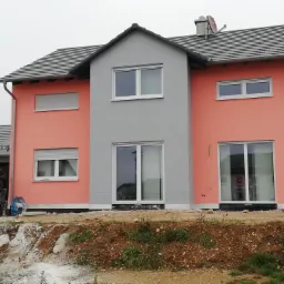 Nowo wybudowany dom z pomarańczową elewacją i szarym fragmentem centralnym, widoczni dwaj pracownicy budowlani i teren wokół domu w trakcie prac wykończeniowych.