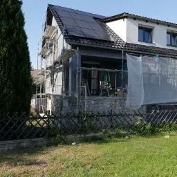 Dom w trakcie ocieplania elewacji: widoczne rusztowania, szary styropian na ścianach, częściowo osłonięty siatką ochronną, z panelami słonecznymi na dachu i kamienną podmurówką.