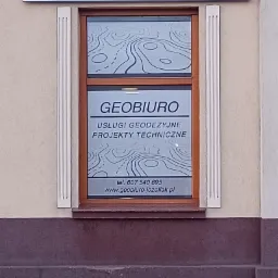 Fasada budynku z szyldem 'GEODEZJA' i oknem z napisem 'GEOBIURO Usługi geodezyjne Projekty techniczne', w oknie widoczny motyw mapy topograficznej.