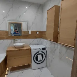 Nowoczesna łazienka z marmurowymi płytkami, drewnianymi szafkami, umywalką nablatową i pralką z zawieszką informacyjną, odbicie osoby robiącej zdjęcie w lustrze.