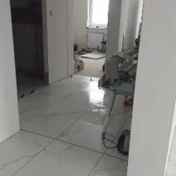 Ułożone białe płytki podłogowe z szarym marmurkowym wzorem, widoczna przecinarka do płytek, w tle pomieszczenie w trakcie prac wykończeniowych z oknem.