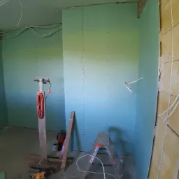 Wnętrze w trakcie montażu zielonych płyt kartonowo-gipsowych, widoczne narzędzia, drabina aluminiowa, oświetlenie robocze i przewody elektryczne pod sufitem.