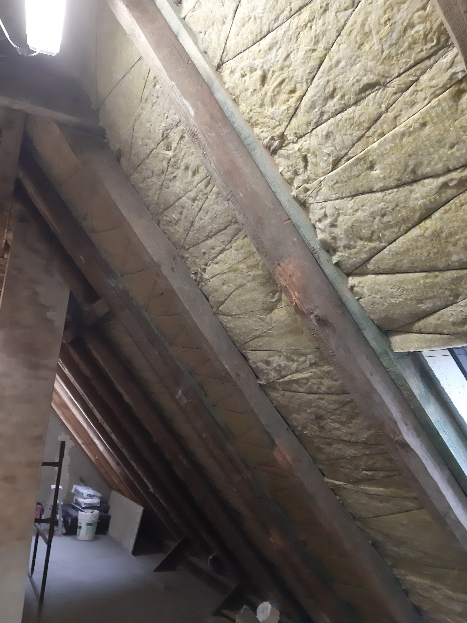 Izolacja poddasza wełną mineralną pomiędzy krokwiami, widoczne drewniane elementy konstrukcyjne dachu i częściowo wykończona ściana.