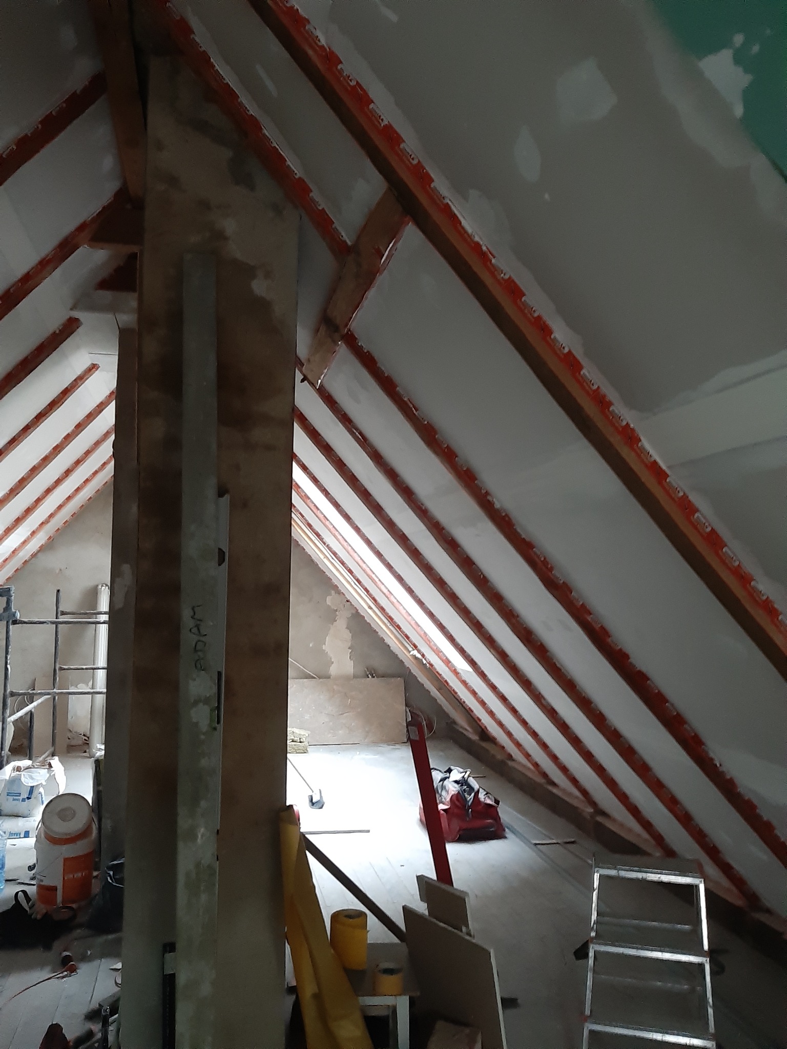 Poddasze w trakcie adaptacji: widoczne drewniane krokwie z zamontowanymi profilami, częściowo pokryte płytami gipsowo-kartonowymi, surowe ściany, narzędzia i materiały budowlane porozrzucane...