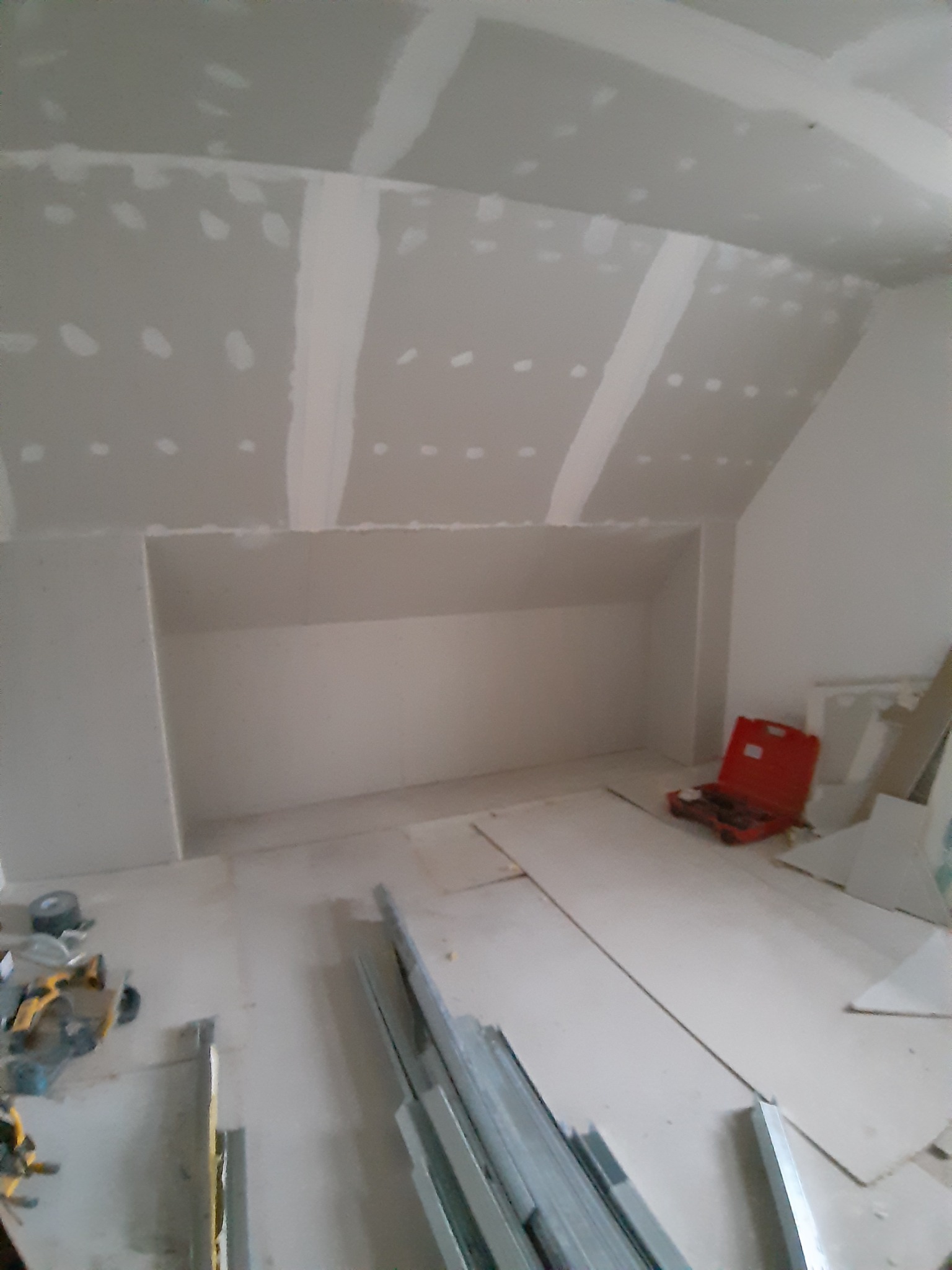 Poddasze ze skośnym sufitem w trakcie montażu płyt kartonowo-gipsowych, widoczna wnęka, narzędzia i profile na podłodze, szpachlowane łączenia płyt.