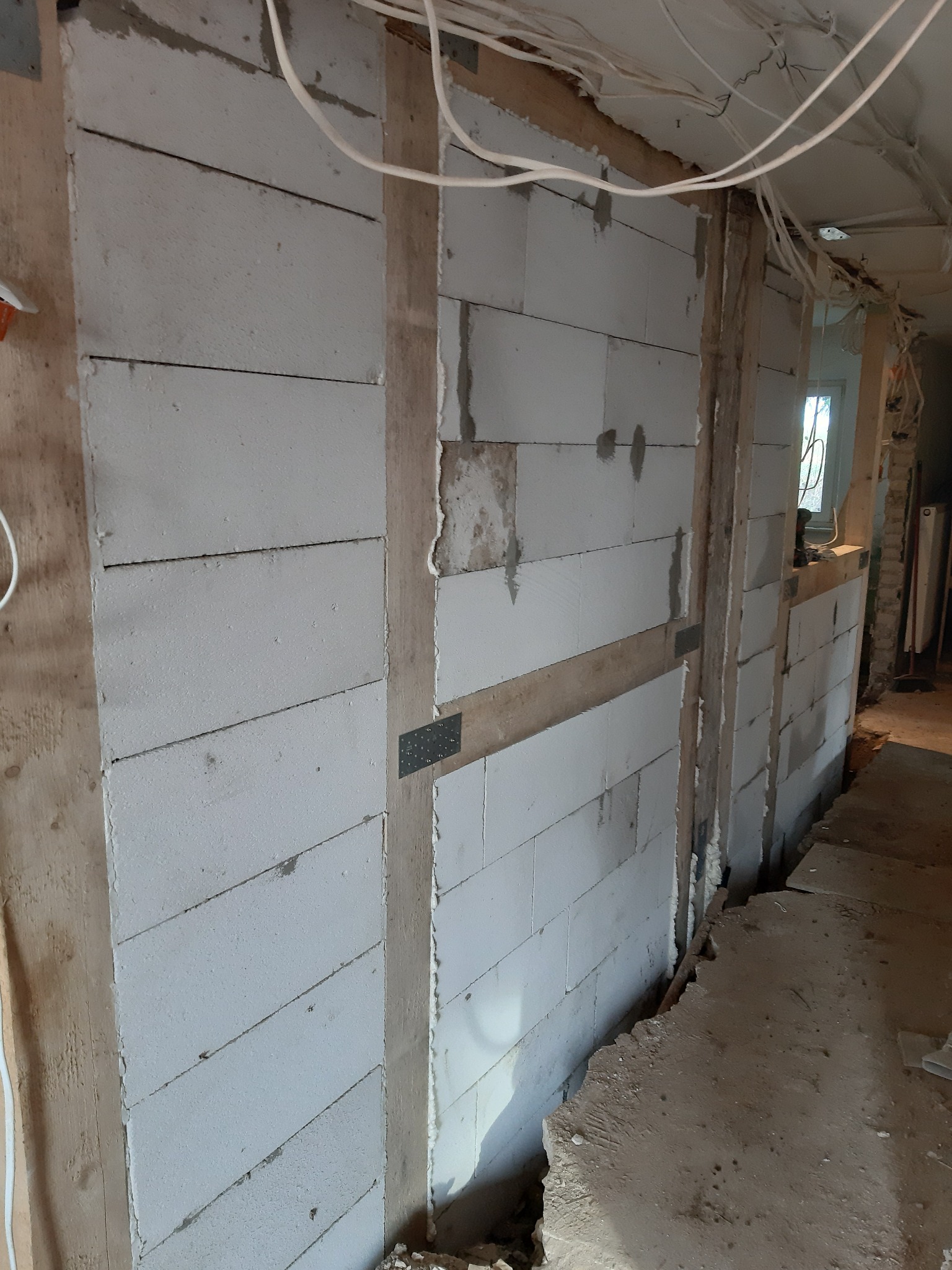 Wznoszenie ściany z białych bloczków, widoczne drewniane wzmocnienia pionowe i poziome, metalowe łączniki, wystające przewody elektryczne i gruz na podłodze.