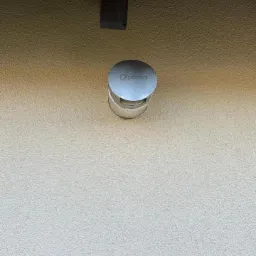 Metalowa czerpnia powietrza z logo na elewacji budynku o jasnej, chropowatej strukturze.