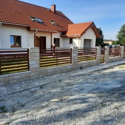 PRZEMYSŁAW BARTNIK STĘBUD - Nowoczesne ogrodzenie posesji z poziomych, brązowych desek i beżowych, kwadratowych słupków, na tle białego domu z czerwoną dachówką i błękitnego nieba.