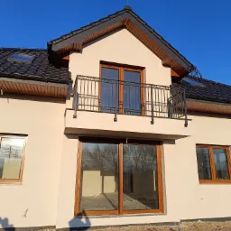 Nowy dom jednorodzinny z balkonem z kutą balustradą, brązowymi oknami i drzwiami balkonowymi, czarną dachówką i beżową elewacją w słoneczny dzień.