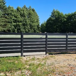 PRZEMYSŁAW BARTNIK STĘBUD - Nowoczesne ogrodzenie panelowe w kolorze czarnym z poziomymi, białymi akcentami, brama wjazdowa. Tło: zieleń drzew i błękitne niebo. Widoczny fragment skrzynki elektrycznej.