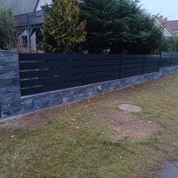 PRZEMYSŁAW BARTNIK STĘBUD - Nowoczesne ogrodzenie panelowe w kolorze antracytowym z podmurówką z szarej kostki, na tle zielonego trawnika i drzew. Widoczny fragment domu w tle.