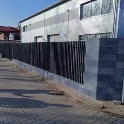 PRZEMYSŁAW BARTNIK STĘBUD - Nowoczesne ogrodzenie z pionowych, czarnych sztachet metalowych, osadzone na szarych, betonowych słupkach i podmurówce. Tło stanowi budynek przemysłowy.