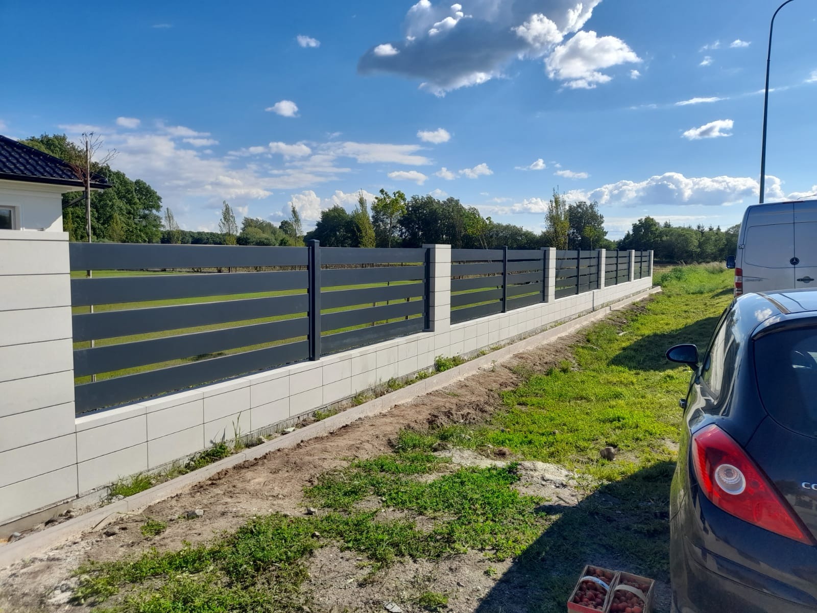 Nowoczesne ogrodzenie panelowe z poziomymi, szarymi przęsłami na betonowej podmurówce, widoczne na tle błękitnego nieba i zieleni trawnika.