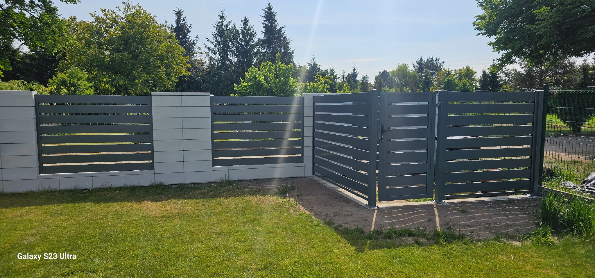 Nowoczesne ogrodzenie z poziomych, szarych paneli i betonowych słupków na tle zielonego trawnika i drzew. Widoczna otwarta furtka.