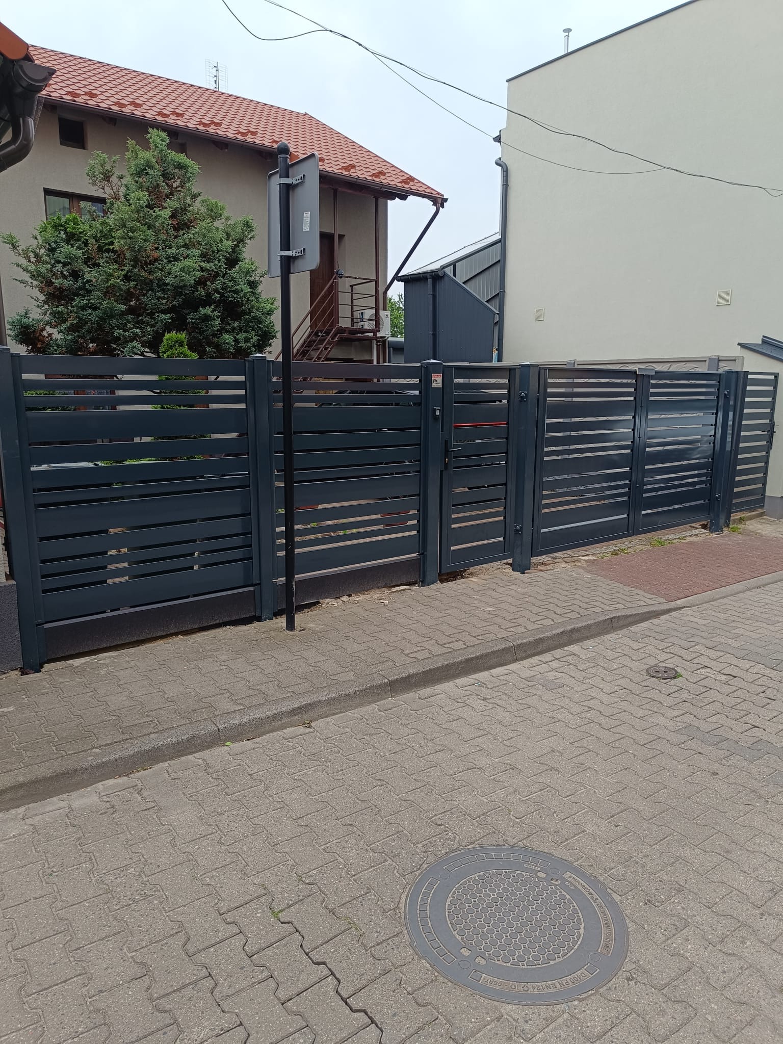 Nowoczesne, poziome ogrodzenie panelowe w kolorze antracytowym, z furtką i przesuwną bramą, na tle domu jednorodzinnego. Widok z poziomu chodnika.