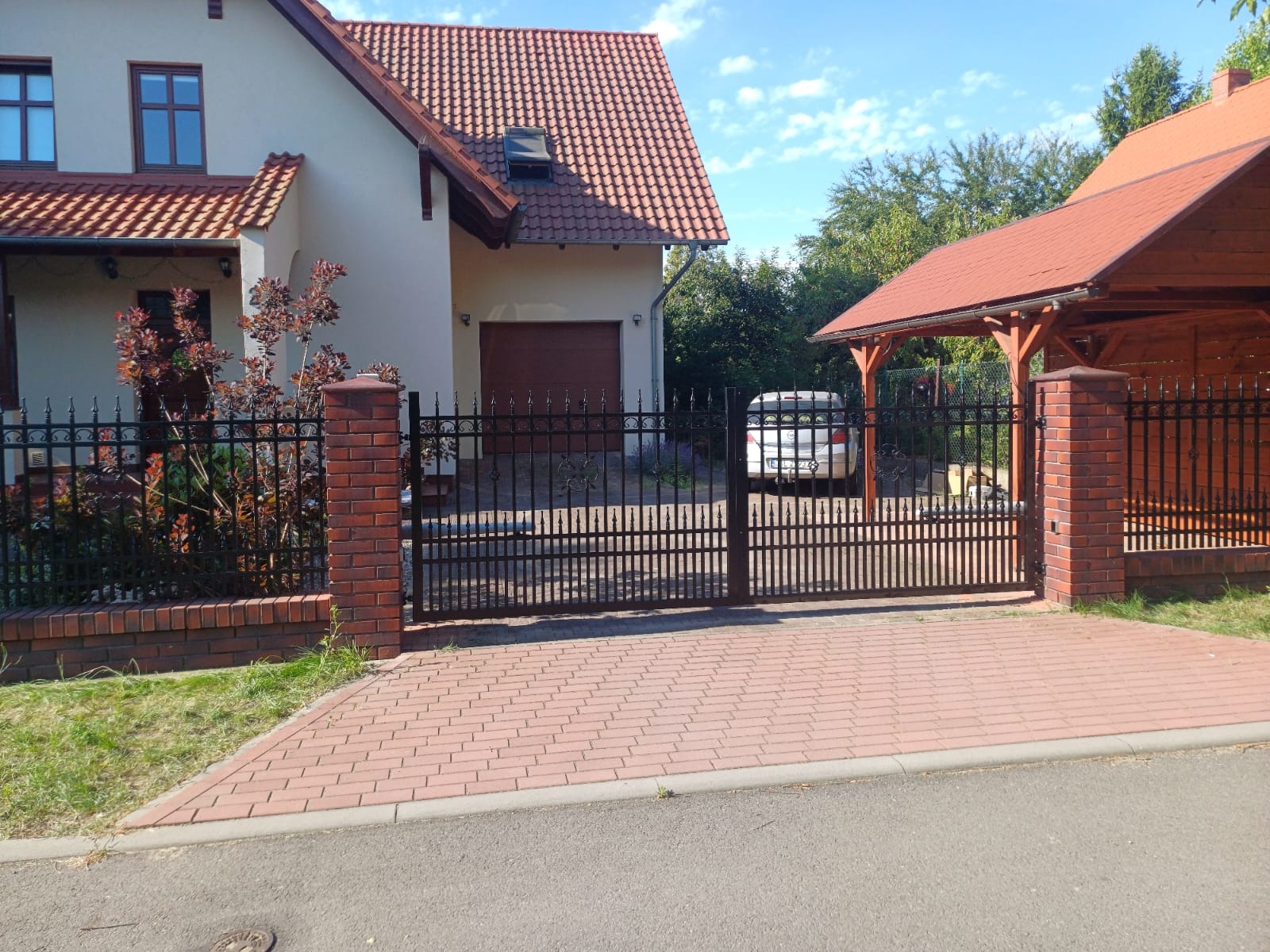 Czarna metalowa brama wjazdowa z dekoracyjnymi elementami, osadzona na ceglanych słupkach, prowadząca do posesji z domem i wiatą garażową.