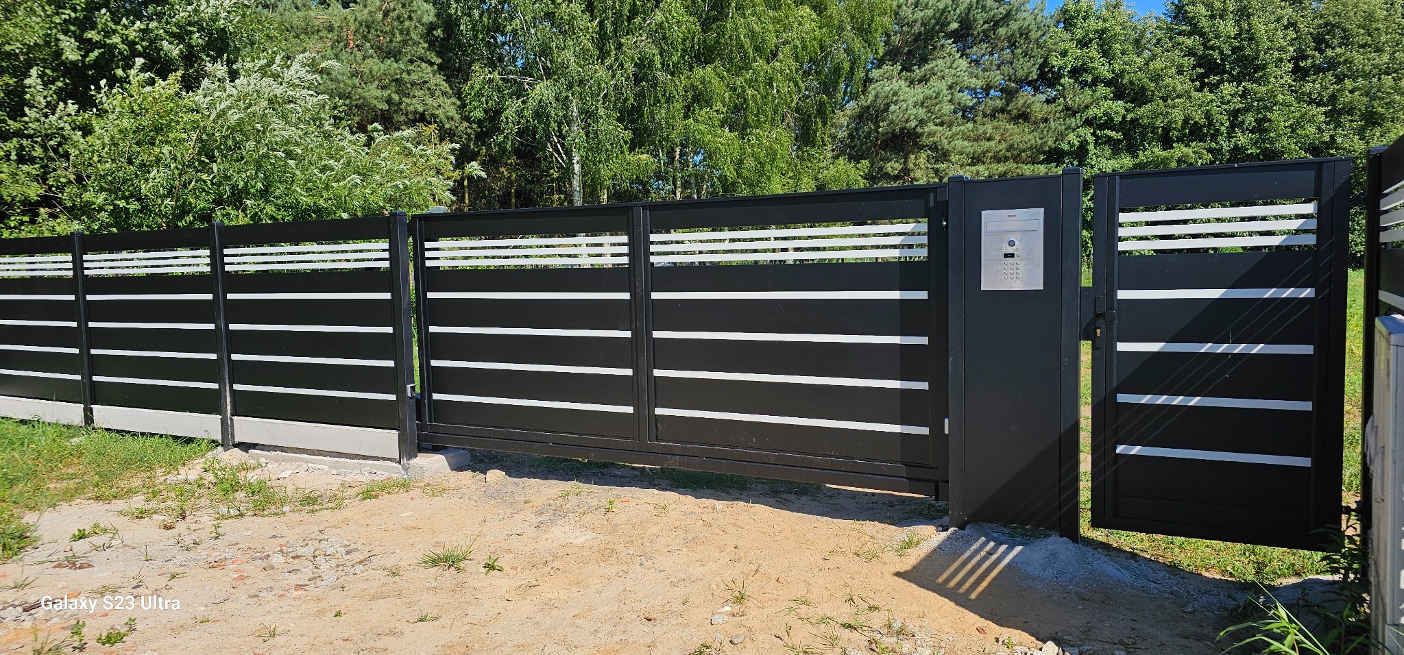 Nowoczesna, przesuwna brama wjazdowa z poziomymi, białymi akcentami na tle zieleni. Metalowa konstrukcja w kolorze antracytu z panelem domofonu.