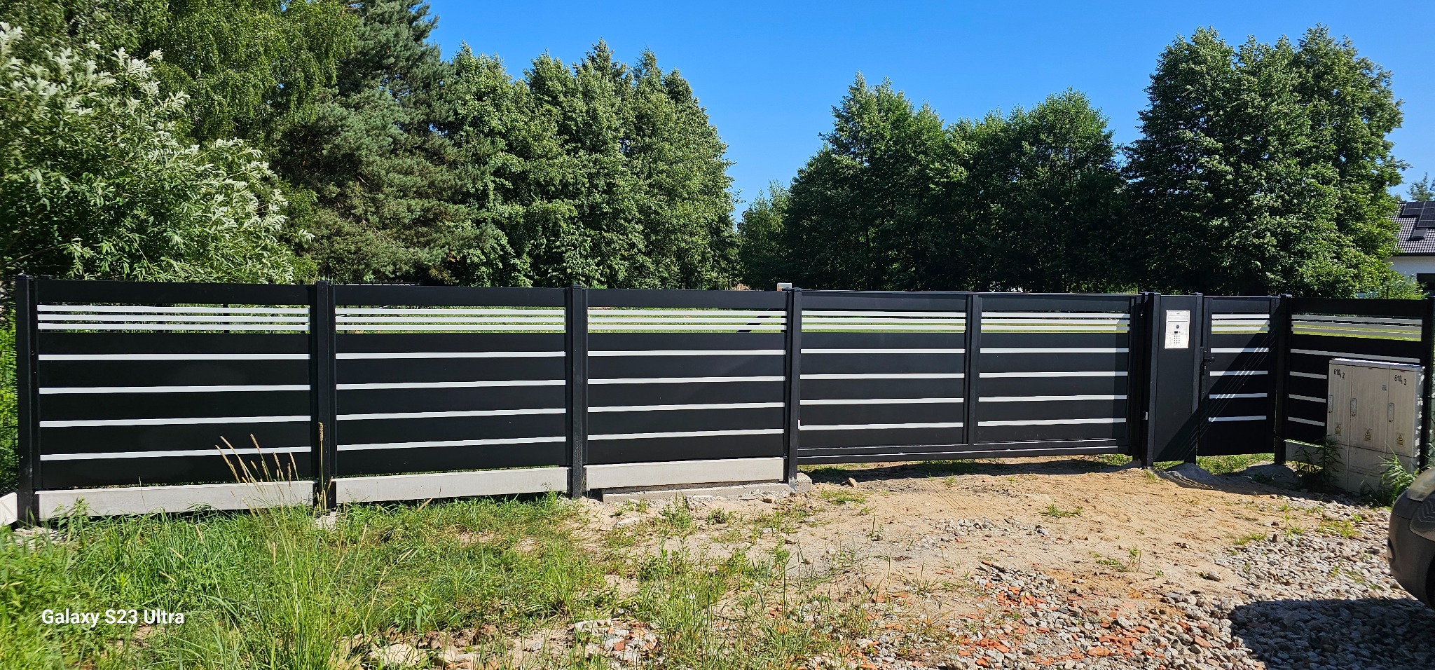 Nowoczesne ogrodzenie panelowe w kolorze czarnym z poziomymi, białymi akcentami, brama wjazdowa. Tło: zieleń drzew i błękitne niebo. Widoczny fragment skrzynki elektrycznej.