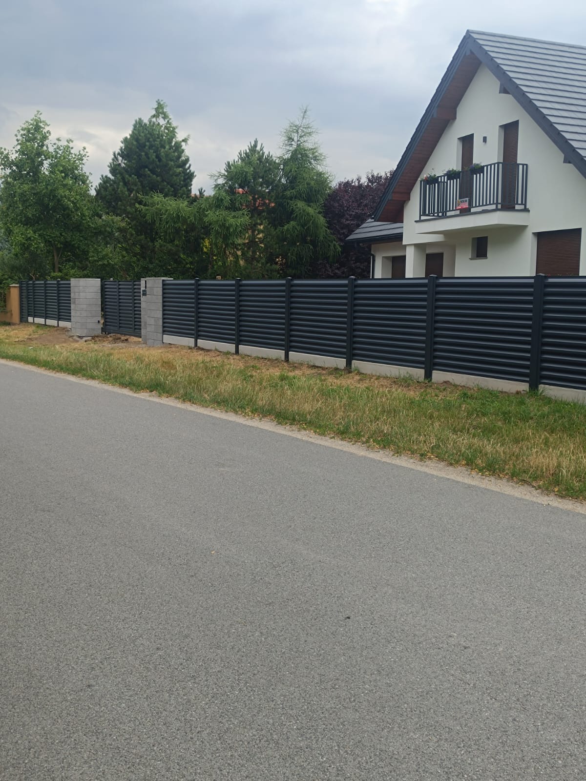 Nowoczesne, poziome ogrodzenie metalowe w kolorze grafitowym, z bramą i słupkami z szarego betonu, na tle domu z balkonem i zielenią.