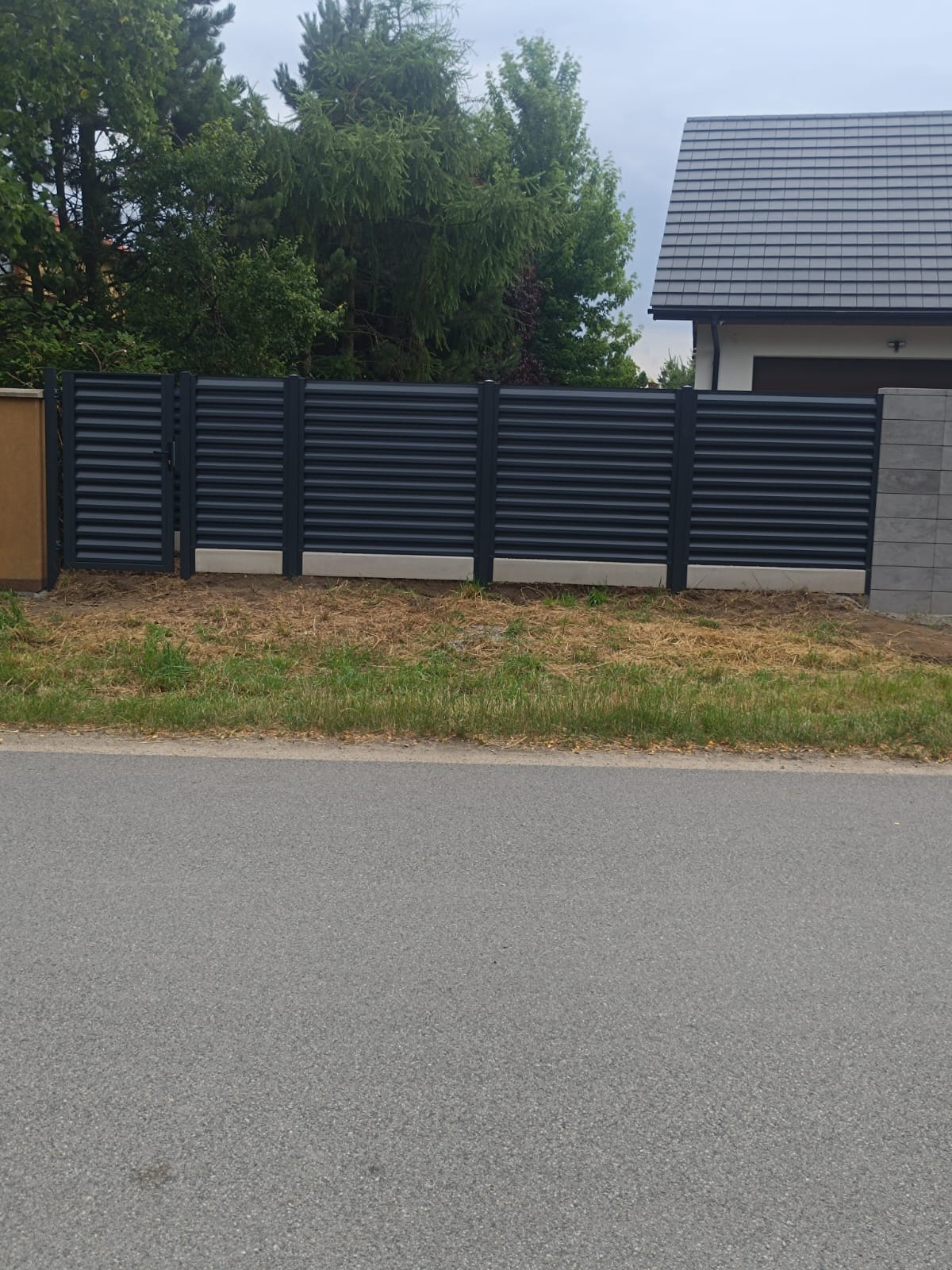 Nowoczesne, poziome ogrodzenie metalowe w kolorze antracytowym, przylegające do asfaltowej drogi. Tło z zielenią drzew i fragmentem domu z szarym dachem.