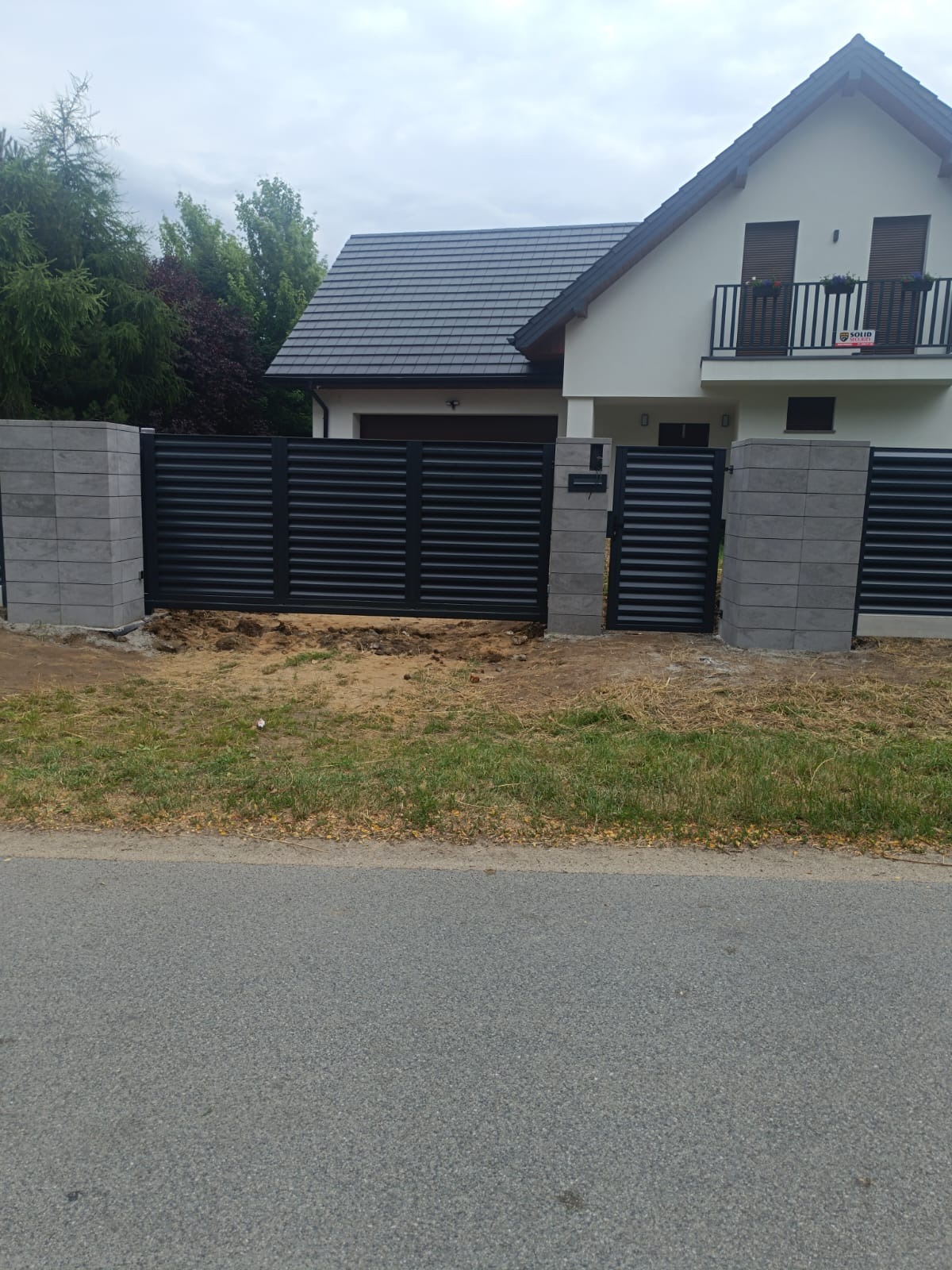 Nowoczesna, pozioma, czarna brama wjazdowa i furtka z szarymi słupkami z bloczków betonowych na tle domu z balkonem.