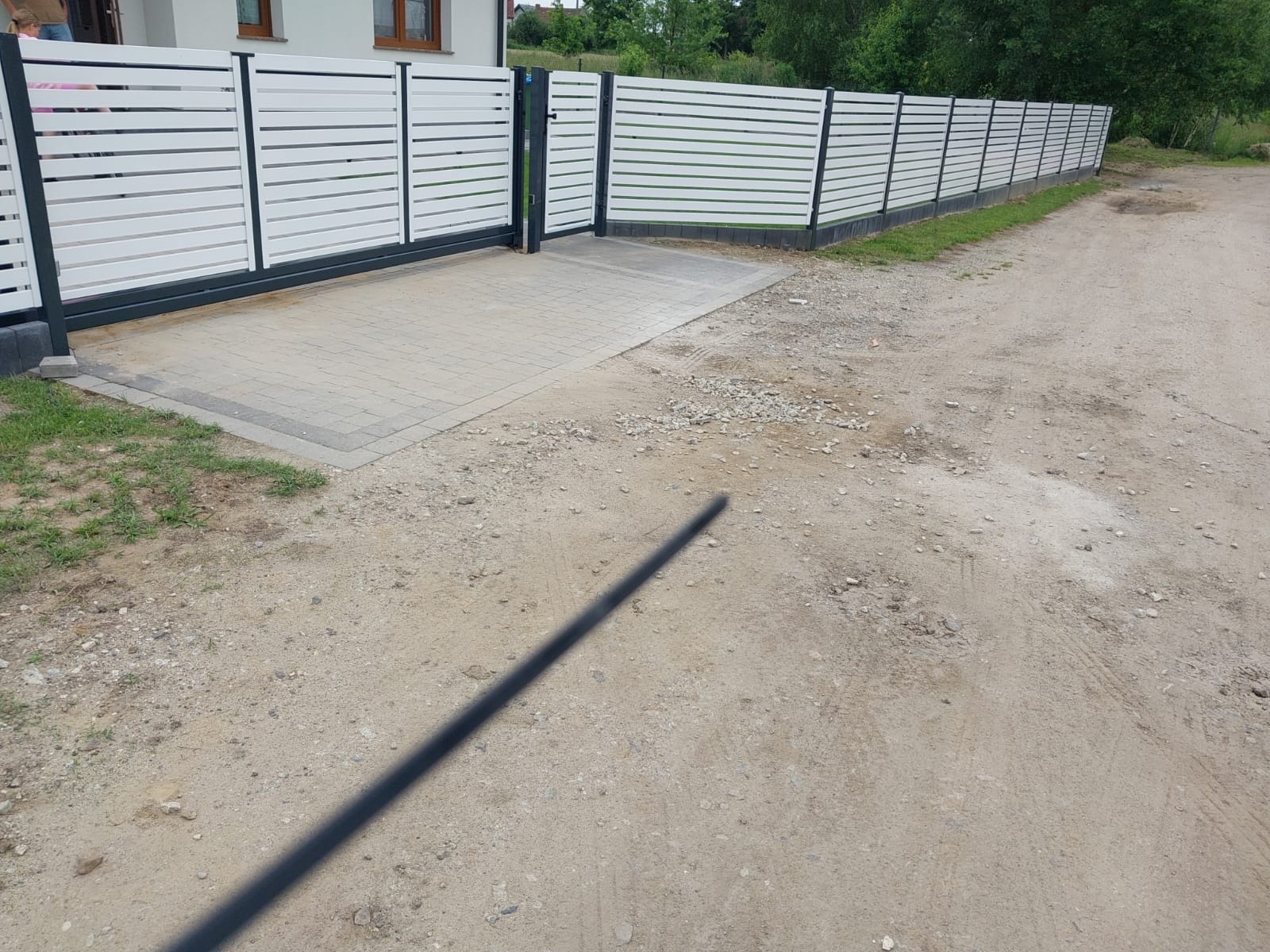 Nowoczesne ogrodzenie panelowe z poziomymi, białymi lamelami i ciemnoszarą ramą, z bramą wjazdową na posesję, widok z zewnątrz na żwirową drogę.
