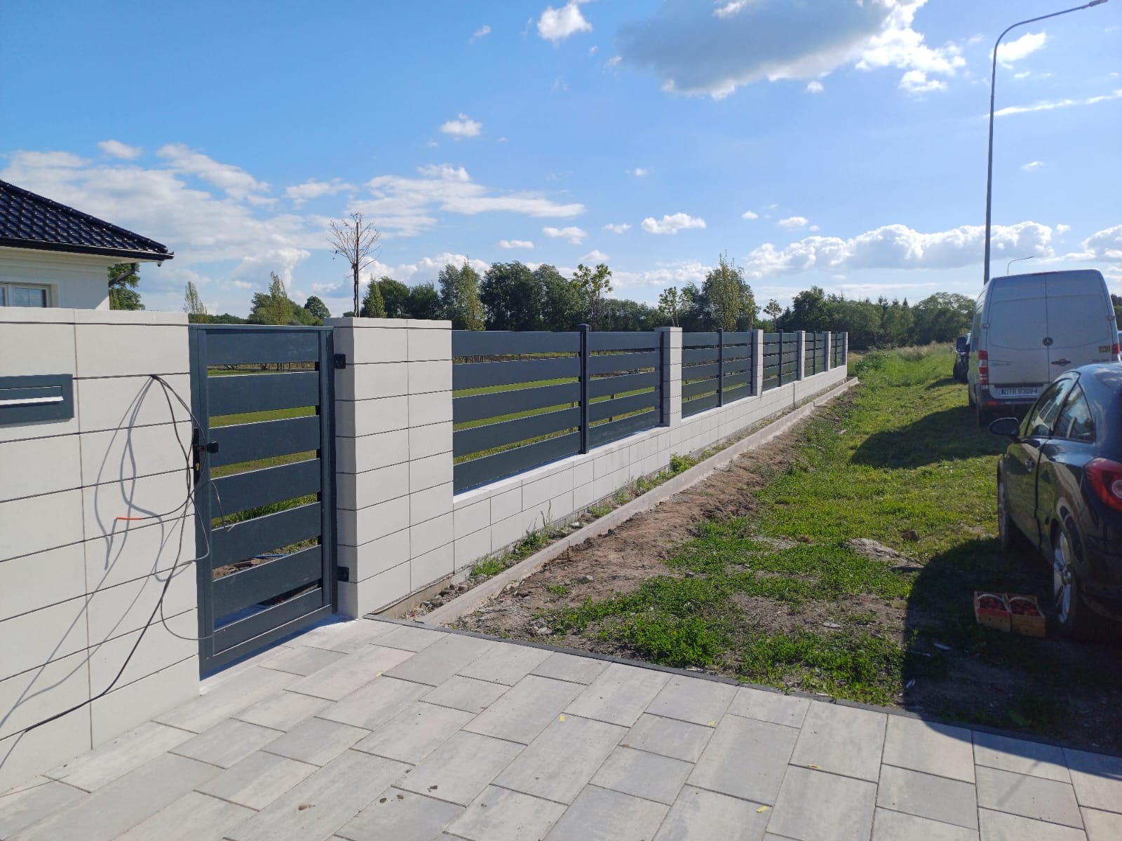 Nowoczesne ogrodzenie z metalowymi przęsłami i betonowymi słupkami, brama wejściowa, kostka brukowa, trawnik, samochody w tle, słoneczny dzień.