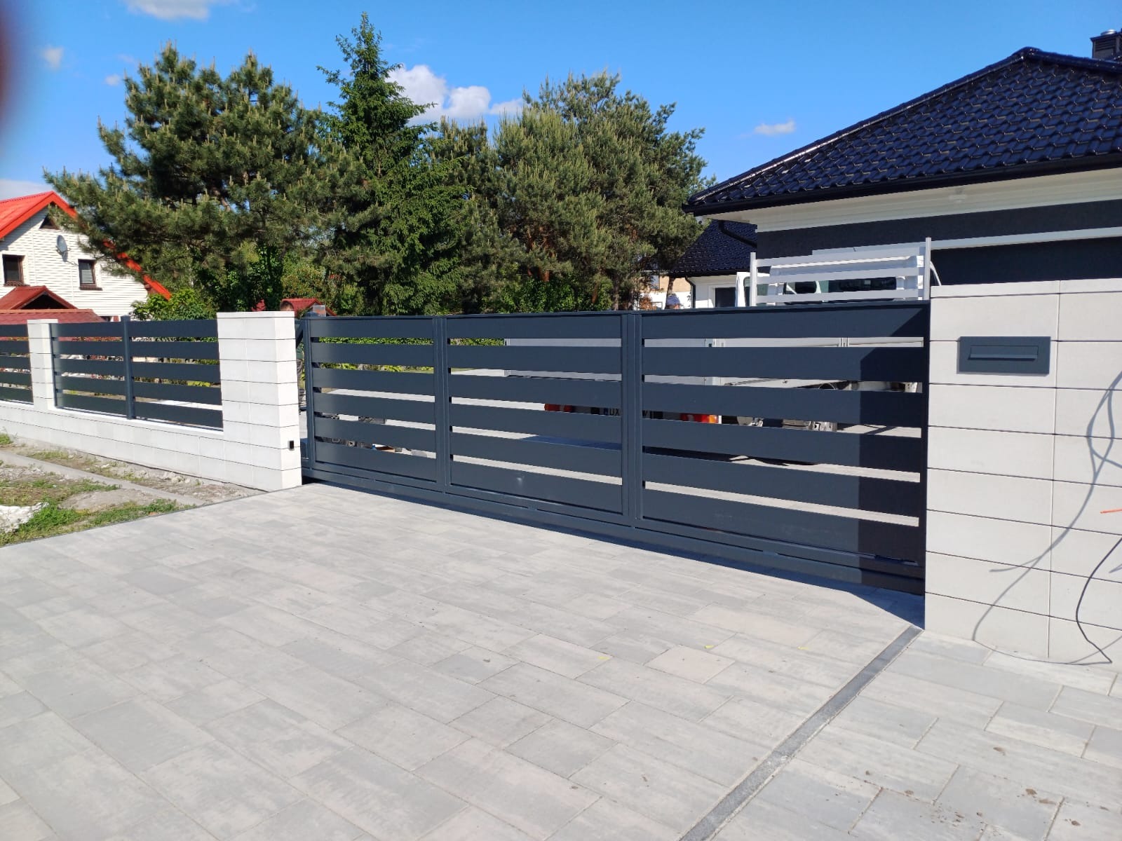 Nowoczesna, przesuwna brama w kolorze antracytowym z poziomymi panelami, zamontowana na posesji z kostką brukową i elementami ogrodzenia z betonu architektonicznego.