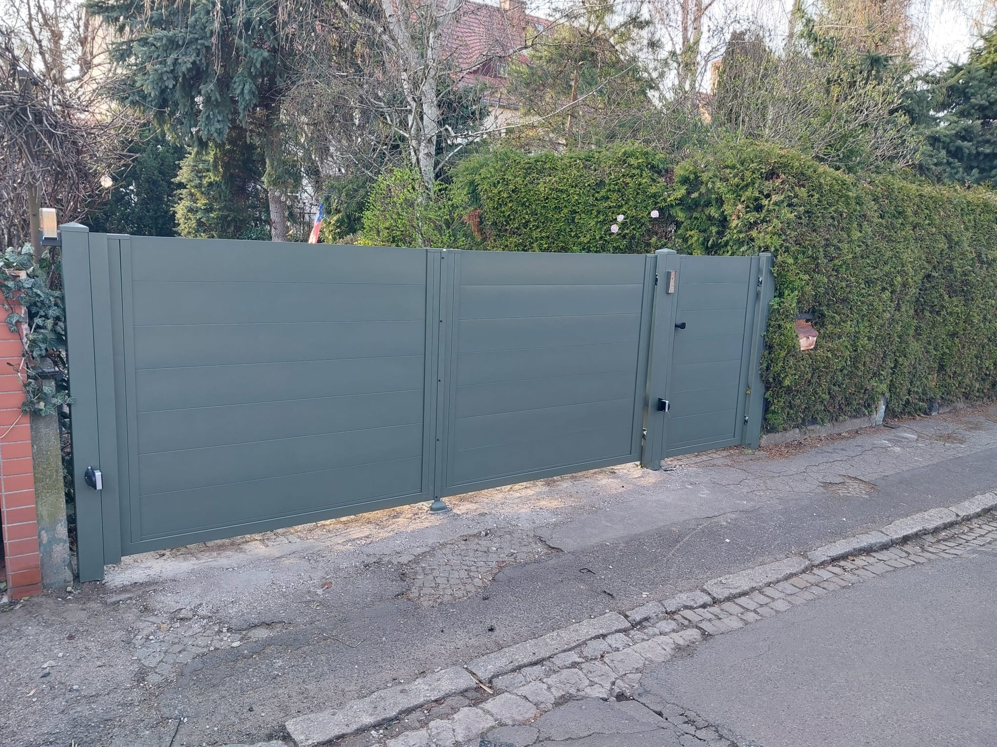 Nowoczesna, aluminiowa brama wjazdowa w kolorze grafitowym, z widocznymi poziomymi panelami, zamontowana przy zielonym żywopłocie i ceglanym słupku.