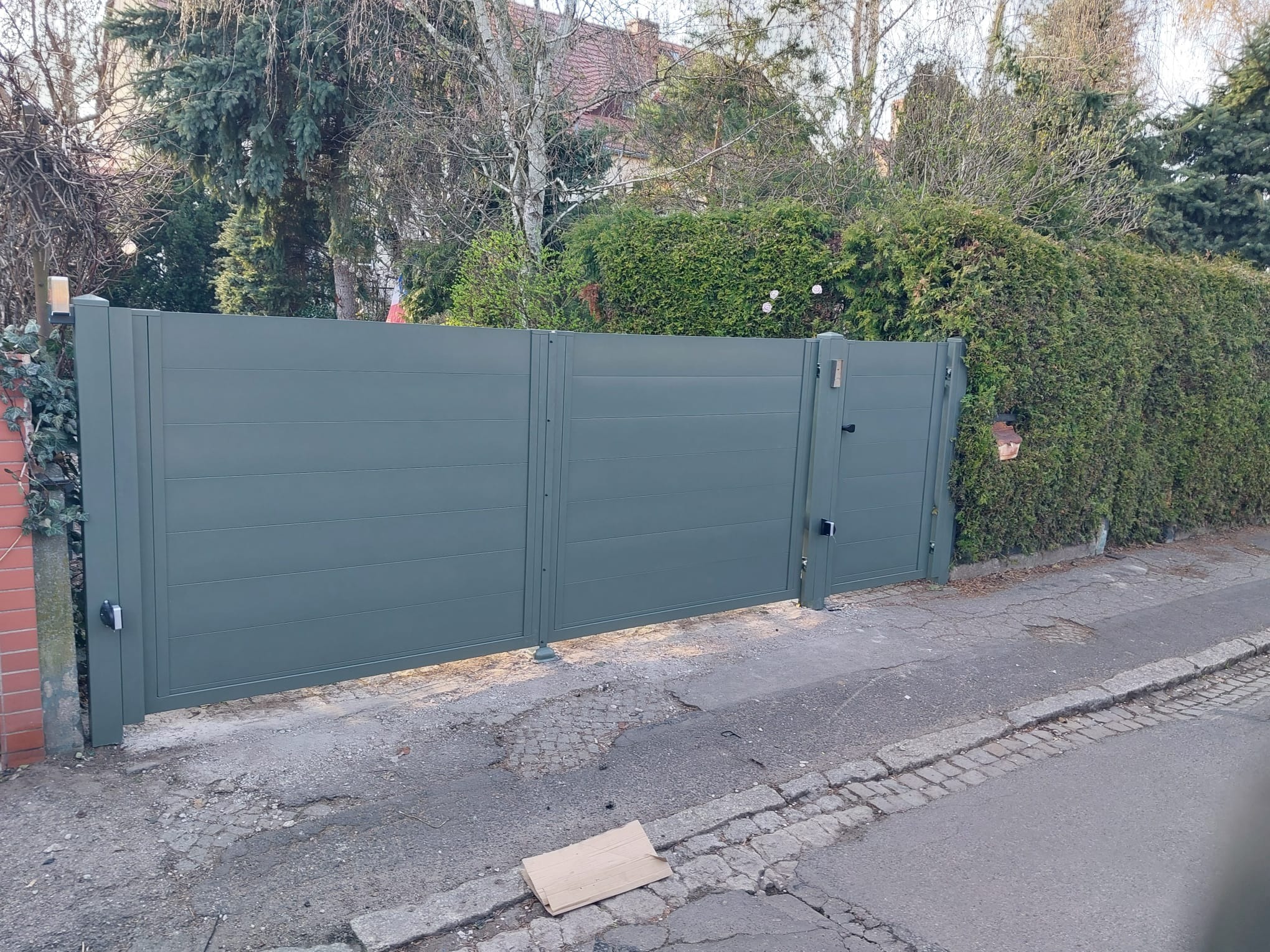 Nowoczesna, dwuskrzydłowa brama w kolorze szarozielonym, z poziomymi panelami, zamontowana na posesji z zielonym żywopłotem i drzewami w tle.