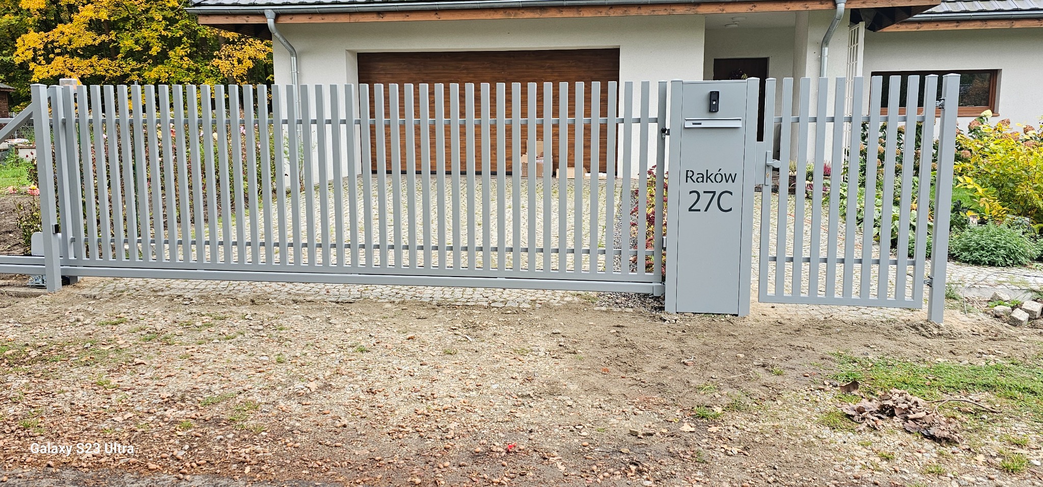 Nowoczesna, przesuwna brama wjazdowa z pionowych, metalowych profili w szarym kolorze. Brama zintegrowana ze skrzynką na listy i domofonem przy domu z garażem.