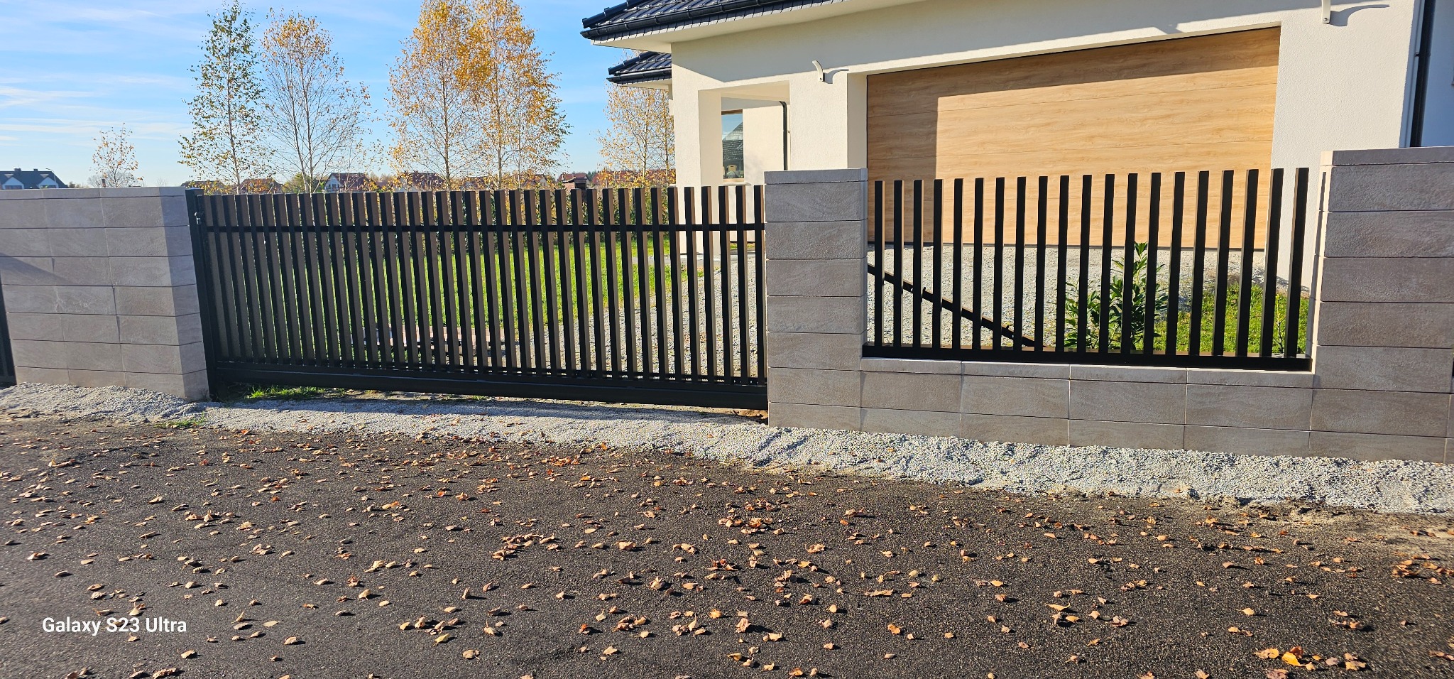 Nowoczesna, przesuwna brama wjazdowa z czarnymi, pionowymi sztachetami, osadzona na niskim murku z beżowych bloczków. Tło: dom z garażem i jesienne drzewa.