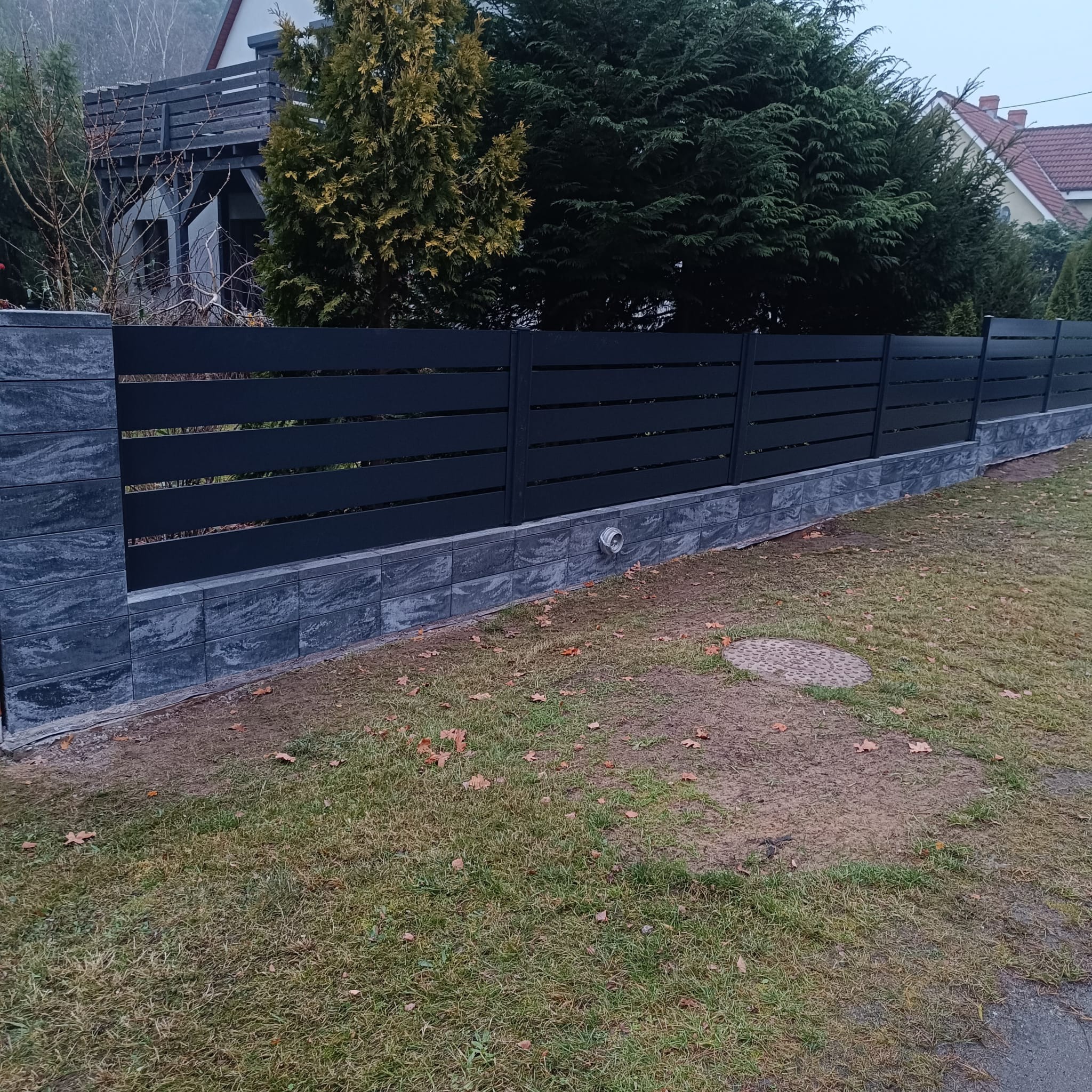 Nowoczesne ogrodzenie panelowe w kolorze antracytowym z podmurówką z szarej kostki, na tle zielonego trawnika i drzew. Widoczny fragment domu w tle.