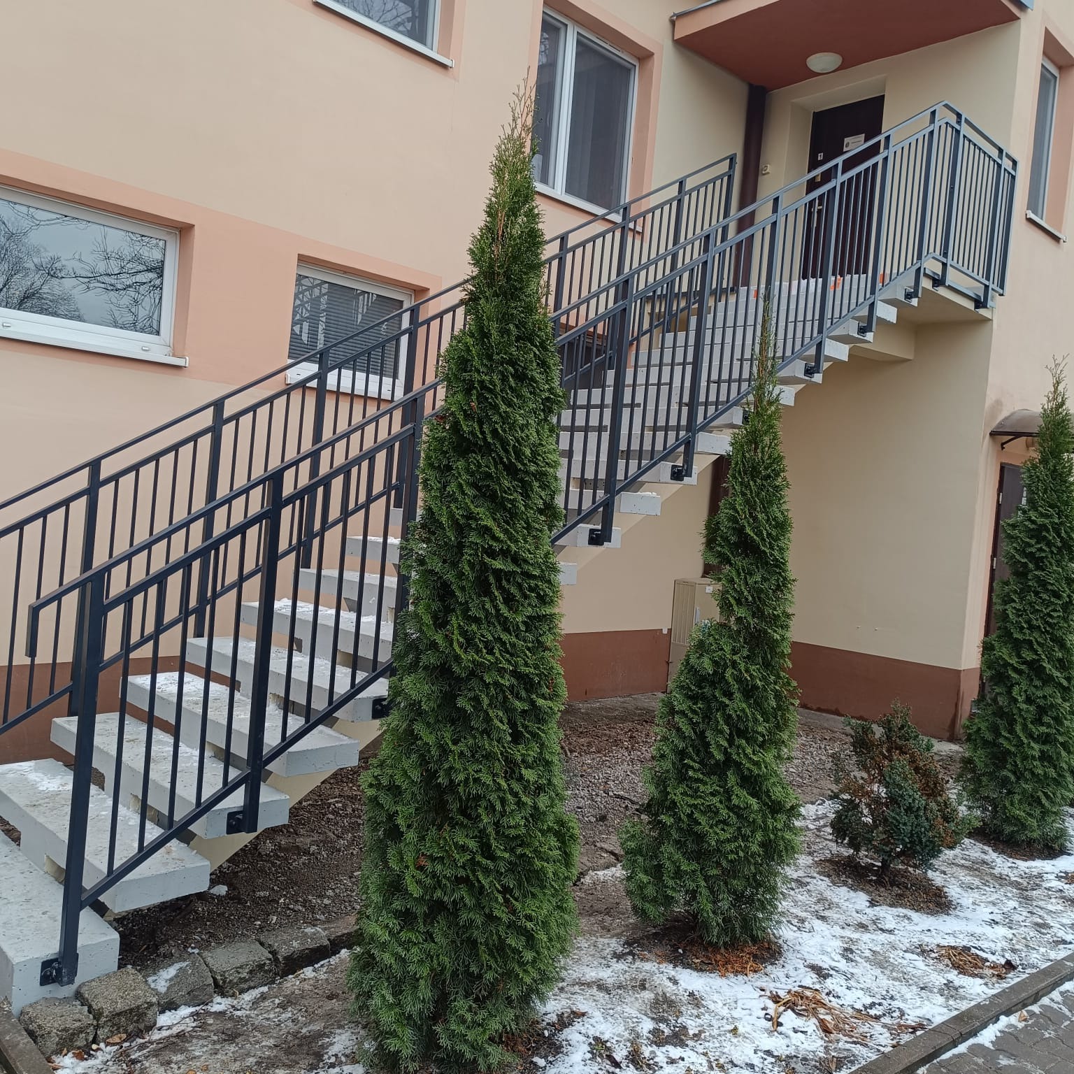 Zewnętrzne schody z czarną metalową balustradą przy budynku, otoczone zielonymi tujami. Widoczne fragmenty śniegu na ziemi. Ujęcie z dołu, podkreślające konstrukcję.