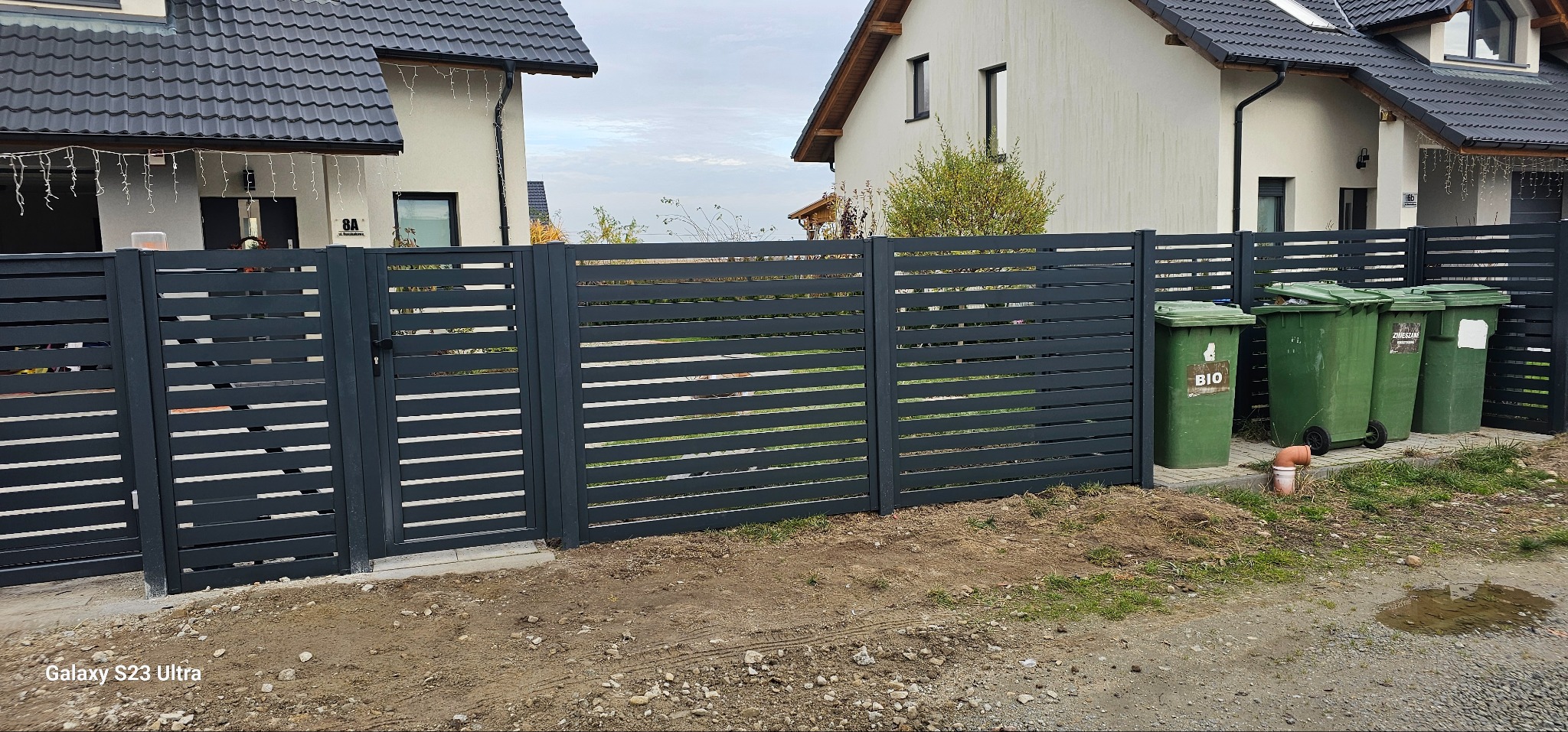 Nowoczesne, poziome ogrodzenie metalowe w kolorze grafitowym z furtką, na tle domów jednorodzinnych i zielonych pojemników na śmieci. Widok z poziomu gruntu.