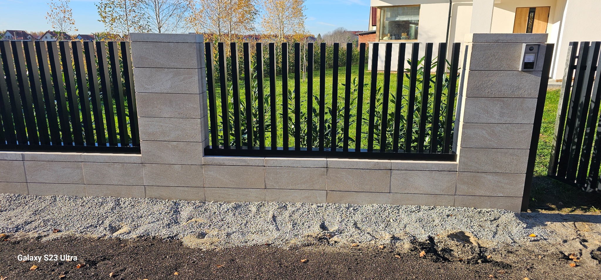 Nowoczesne ogrodzenie z metalowymi sztachetami i kamiennymi słupkami, z wideodomofonem na jednym ze słupków. Trawa widoczna przez szczeliny.