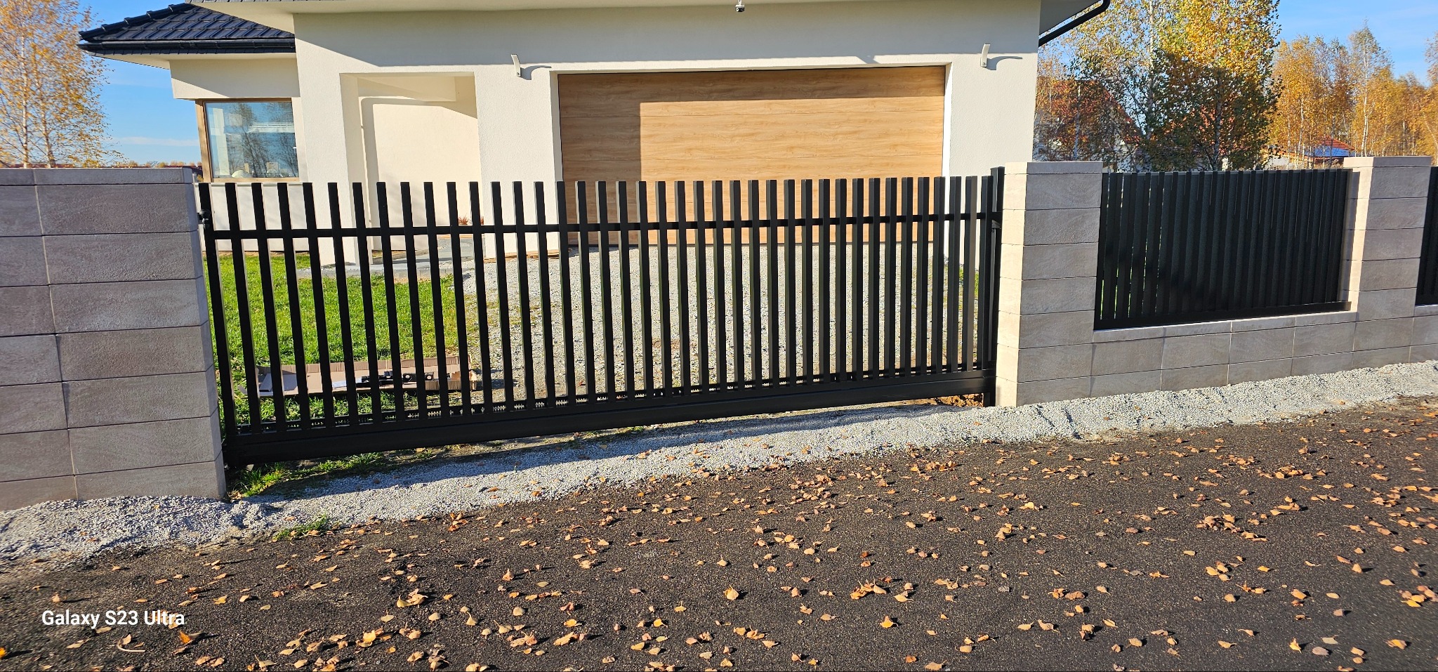 Nowoczesna, przesuwna brama metalowa w kolorze czarnym, z pionowymi szczebelkami, zamontowana między słupkami z jasnych, prostokątnych bloczków betonowych. Tło: dom z garażem.