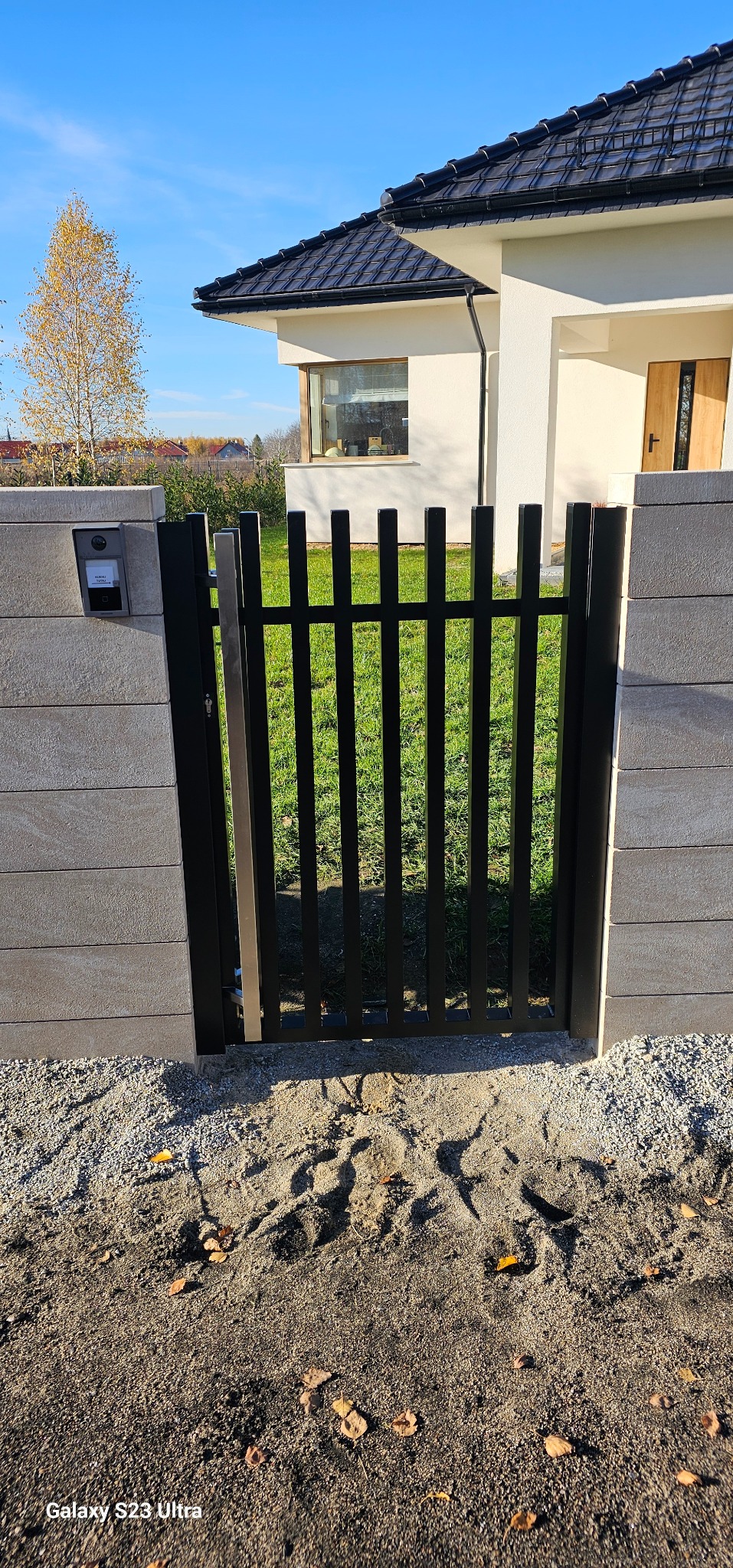 Nowoczesna furtka z metalowymi sztachetami, wkomponowana w betonowe przęsła ogrodzenia. Widoczny dom z jasną elewacją i zielony trawnik w tle. Słoneczny dzień.