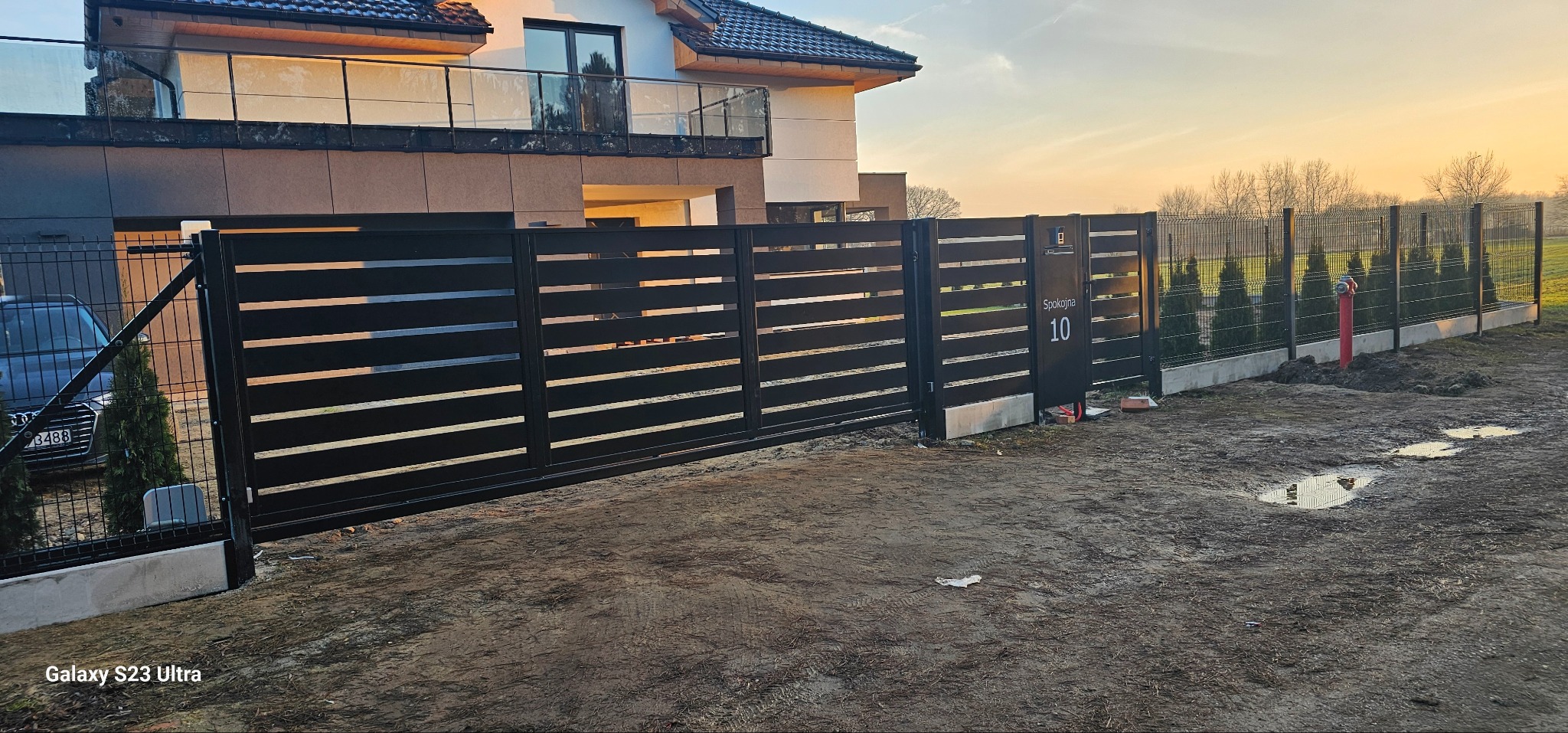 Nowoczesna, przesuwna brama w kolorze antracytu z poziomymi panelami, z numerem domu '10' i skrzynką na listy. W tle dom z balkonem i ogrodzenie panelowe.