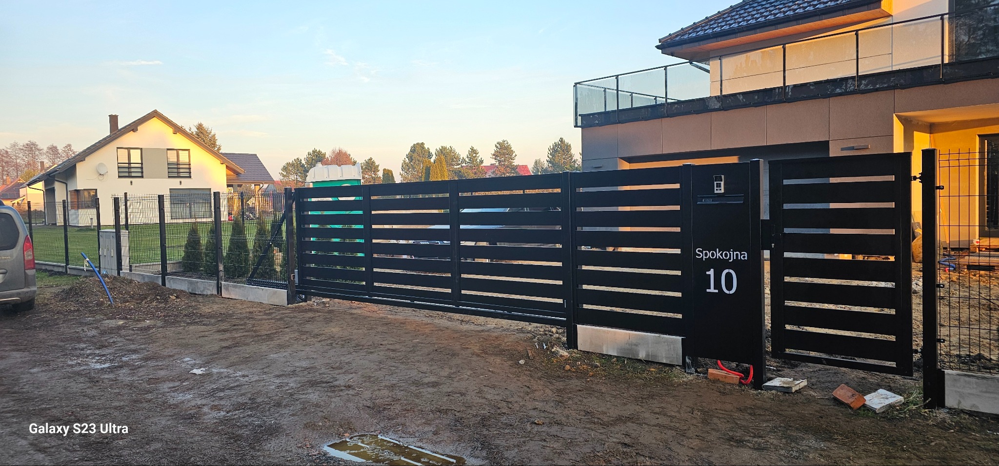 Nowoczesna, przesuwna brama w kolorze czarnym z poziomymi szczeblami, z numerem domu 'Spokojna 10', na tle domu z balkonem i ogrodzenia z siatki.