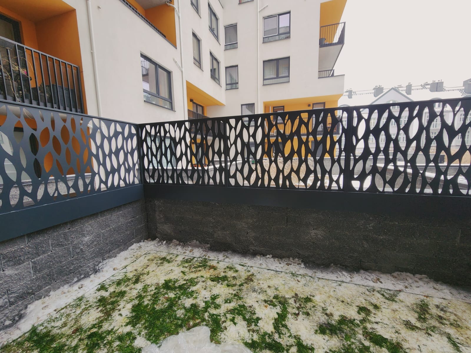 Nowoczesne ogrodzenie metalowe z ażurowym wzorem liści na tle budynku mieszkalnego. Widoczne elementy konstrukcyjne i wykończeniowe. Zastosowano szary kolor.